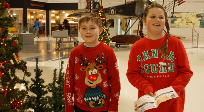 Children wearing Christmas jumpers walking by Christmas trees, holding the museum Christmas trail in their hands.