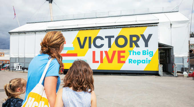 A mother and two children look at a scaffolding tent covering a ship that reads 'Victory Live: The Big Repair'.