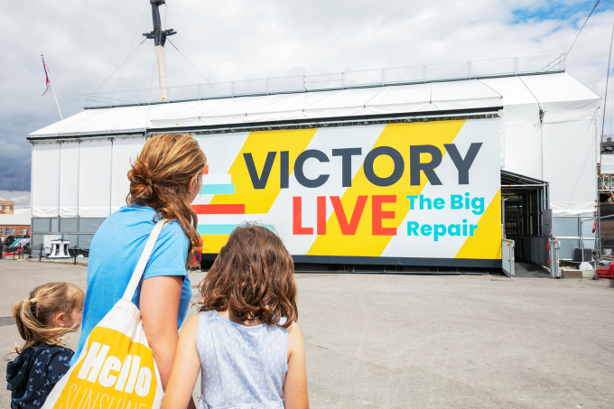 A mother and two young children stand in front of a large sign that says 'Victory Live The Big Repair'