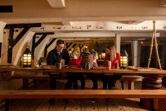 Family sit at a mess table inside HMS Trincomalee, an 18th century tail sail wooden ship of the Royal Navy. 