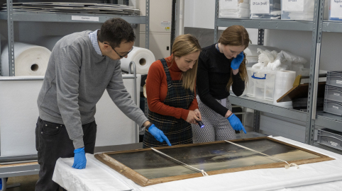 Three people stand looking at a painting on a table