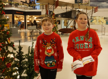 Children wearing Christmas jumpers walking by Christmas trees, holding the museum Christmas trail in their hands.