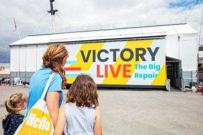 A mother and two children look at a scaffolding tent covering a ship that reads 'Victory Live: The Big Repair'.