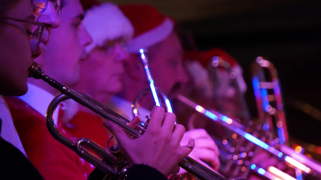 7 brass instrument players playing their instruments, while wearing Christmas hats.