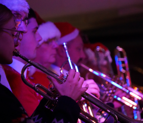 7 brass instrument players playing their instruments, while wearing Christmas hats.