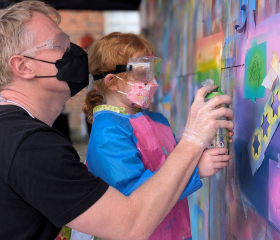 A graffiti artist shows a little girl how to use a spray can on an art wall 