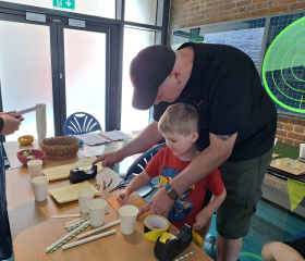 A man and boy look at craft materials laid out across a table.