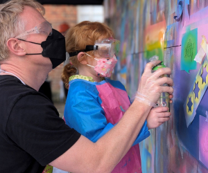 A graffiti artist shows a little girl how to use a spray can on an art wall 