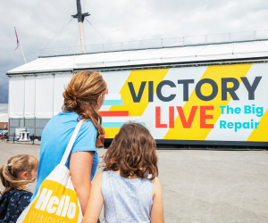 A mother and two children look at a scaffolding tent covering a ship that reads 'Victory Live: The Big Repair'.