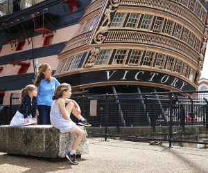 A caregiver and two children sit outside a large wooden ship with a stern bearing the name 'Victory'