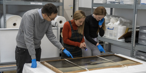 Three people stand looking at a painting on a table