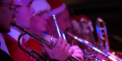 7 brass instrument players playing their instruments, while wearing Christmas hats.