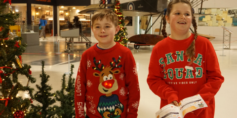 Children wearing Christmas jumpers walking by Christmas trees, holding the museum Christmas trail in their hands.